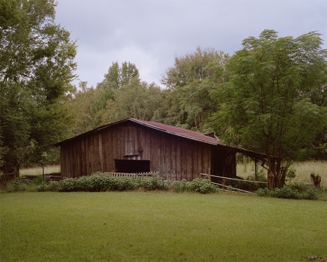Tema Stauffer  Flannery O'Connor's Horse Barn, Andalusia Farm, Milledgeville, Georgia, 2018  Archival Pigment Print  30h x 36w in. a photograph featuring a barn for horses surrounded by trees with a grey cloudy sky