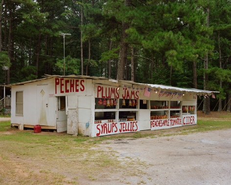 Tema Stauffer  Fruit Stand, Milledgeville Road, Georgia, 2018  Archival Pigment Print  30h x 36w in. a photograph featuring a fruit stand with various fruit names painted on the outer walls, on the side of the road in front of a wall of trees