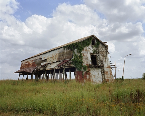 Tema Stauffer  Anderson Cotton Gin, Clarksdale, Mississippi, 2020  Archival Pigment Print  42h x 50 1/2w in. Andersons Cotton Gin Large Rusted steel structure on wooden stilts in a field of greenery with small yellow-orange flowers
